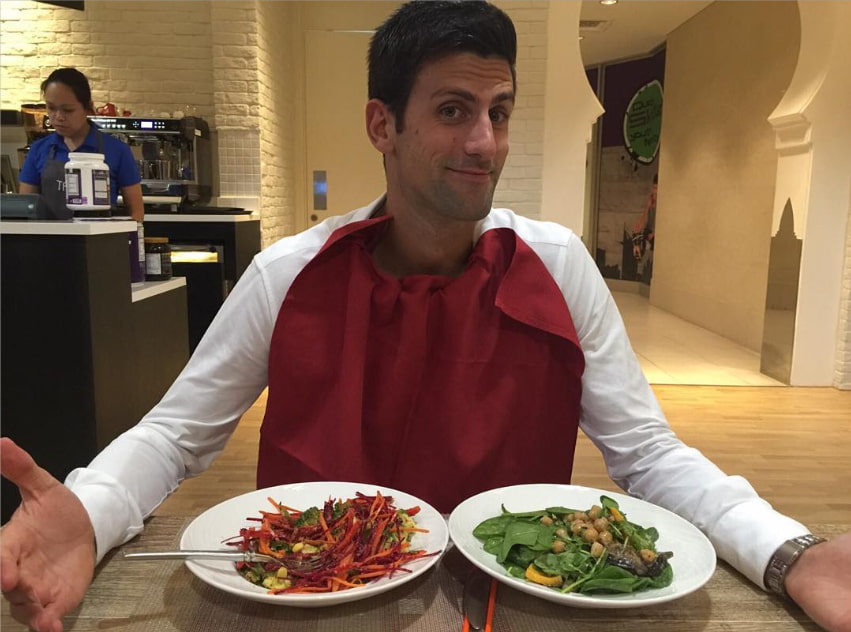Novak Djokovic sonríe y se encoge de hombros frente a dos coloridos bowls de ensalada fresca y sushi, ilustrando su famosa dieta gluten-free y de alto rendimiento en 2026.