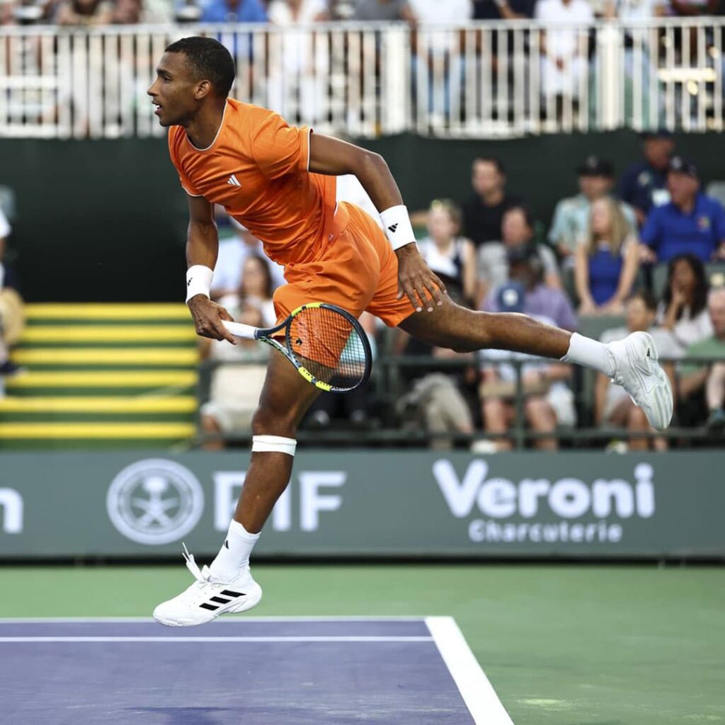 Felix Auger-Aliassime en el aire realizando un estiramiento extremo lateral durante un partido ATP, mostrando la elasticidad de su conjunto Adidas.