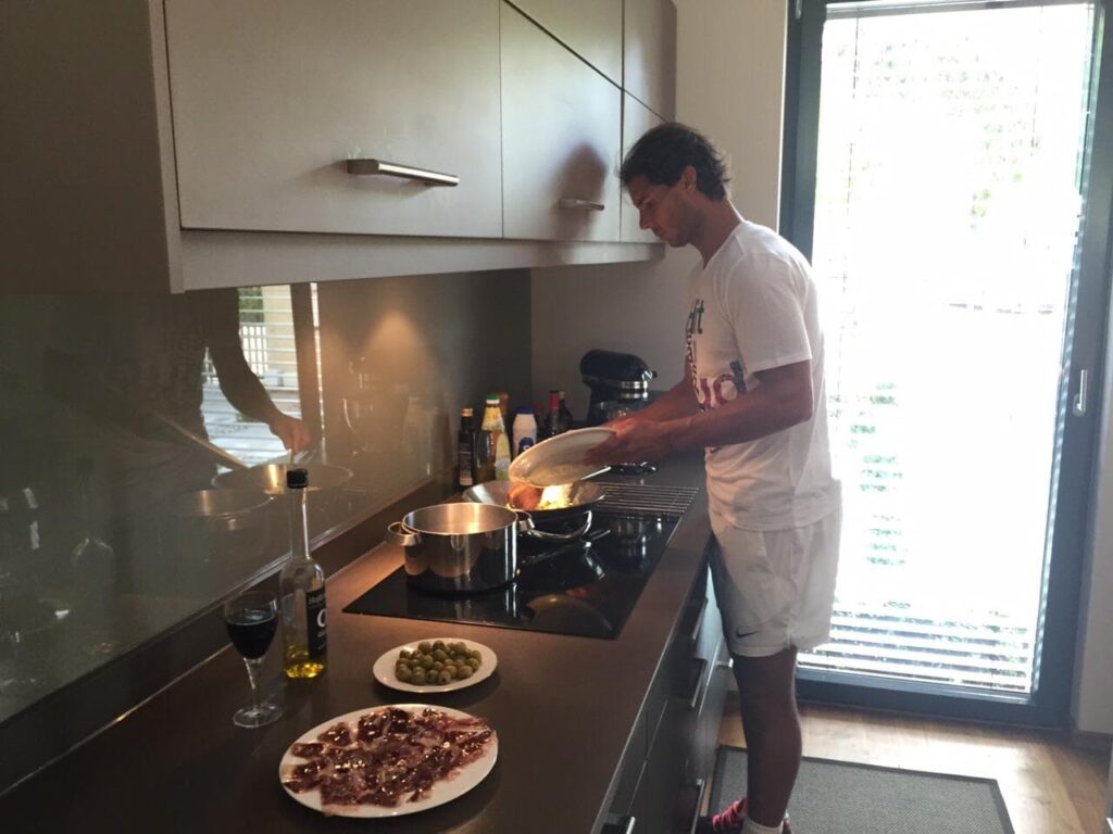 Rafael Nadal concentrado cocinando mariscos en su moderna cocina de Manacor, con jamón ibérico y aceitunas en la encimera.