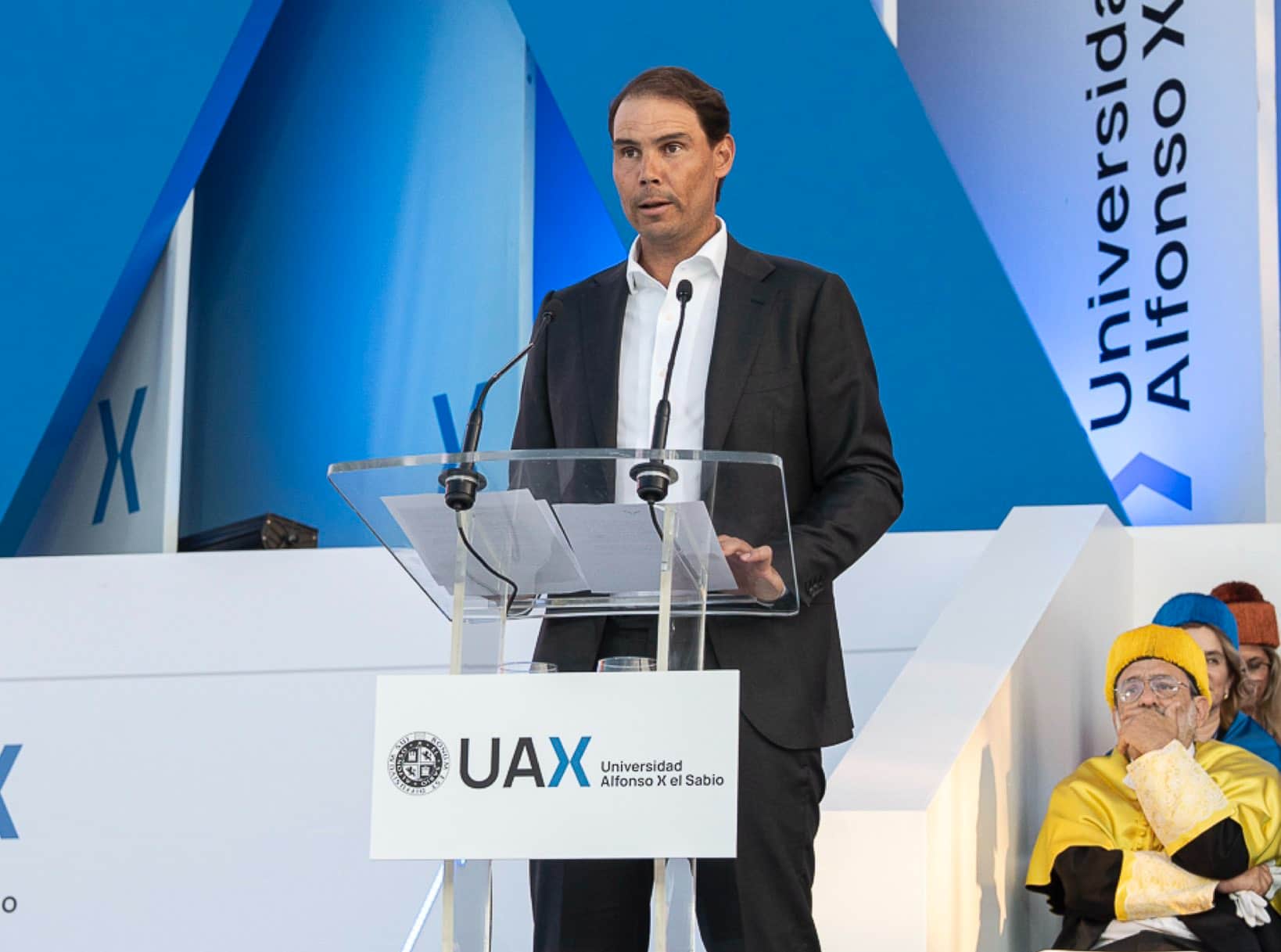 Rafa Nadal con traje oscuro pronunciando un discurso institucional en un podio universitario, representando el rigor y la excelencia analizados en Cancha Central.