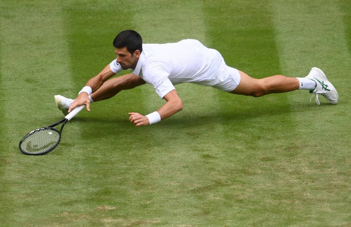 Novak Djokovic realizando un estiramiento extremo tipo yoga para alcanzar una pelota en el césped.