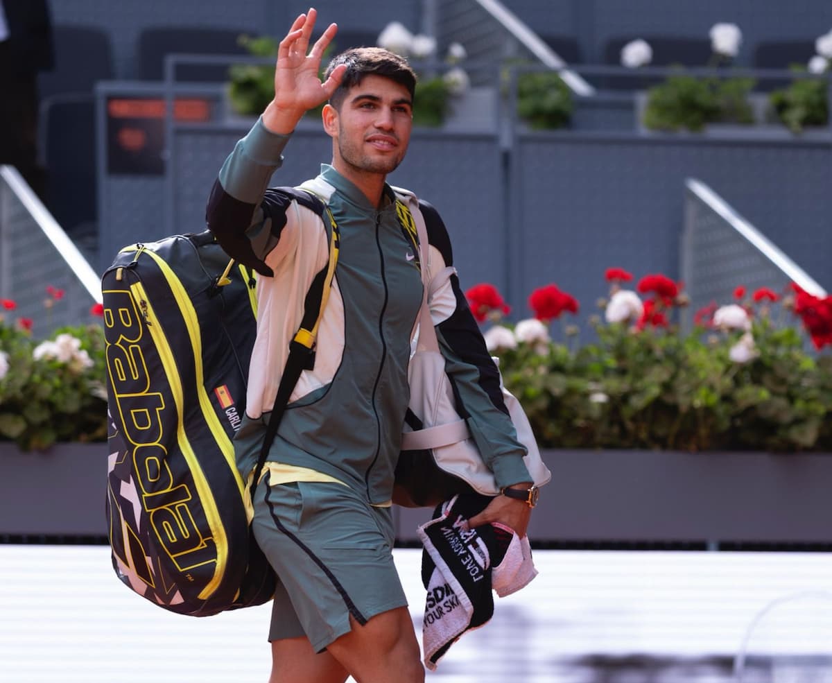 Carlos Alcaraz con indumentaria completa de tenis y raquetero, caminando sobre la tierra batida de la Caja Mágica vacía, levantando la mano en un saludo final de despedida.