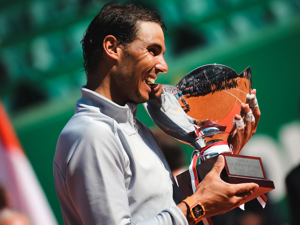 Un Rafael Nadal pletórico y con las manos vendadas, inclinándose para morder el trofeo de campeón en el Masters 1000 de Montecarlo, representando su dominio de 11 títulos en el Principado.