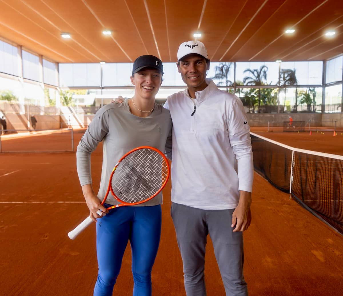 Iga Swiatek ejecutando un revés en las pistas de tierra batida de la Rafa Nadal Academy, con Rafael Nadal observando la sesión desde el fondo.