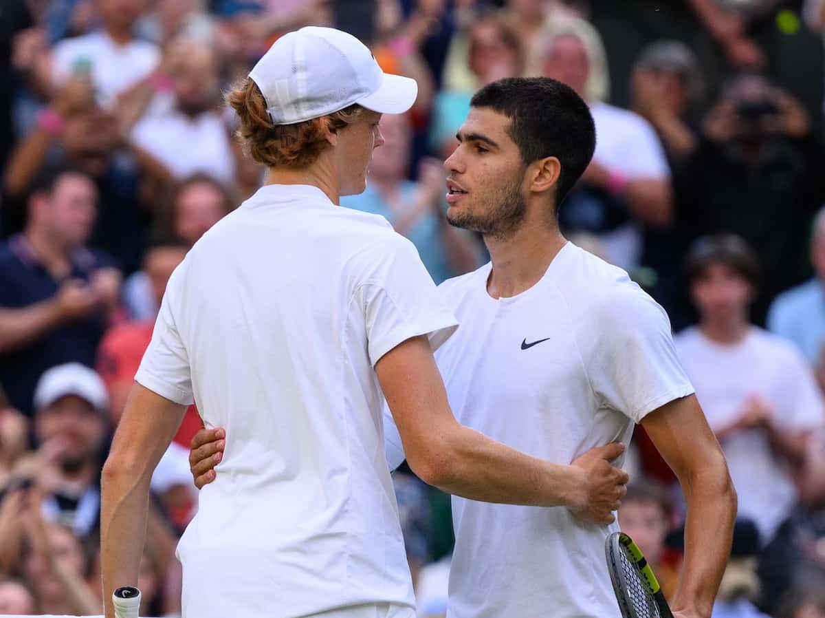 Primer plano de Jannik Sinner (de espaldas, con gorra blanca) y Carlos Alcaraz (de frente) abrazándose en la red tras un partido en Wimbledon, ambos vistiendo de blanco.