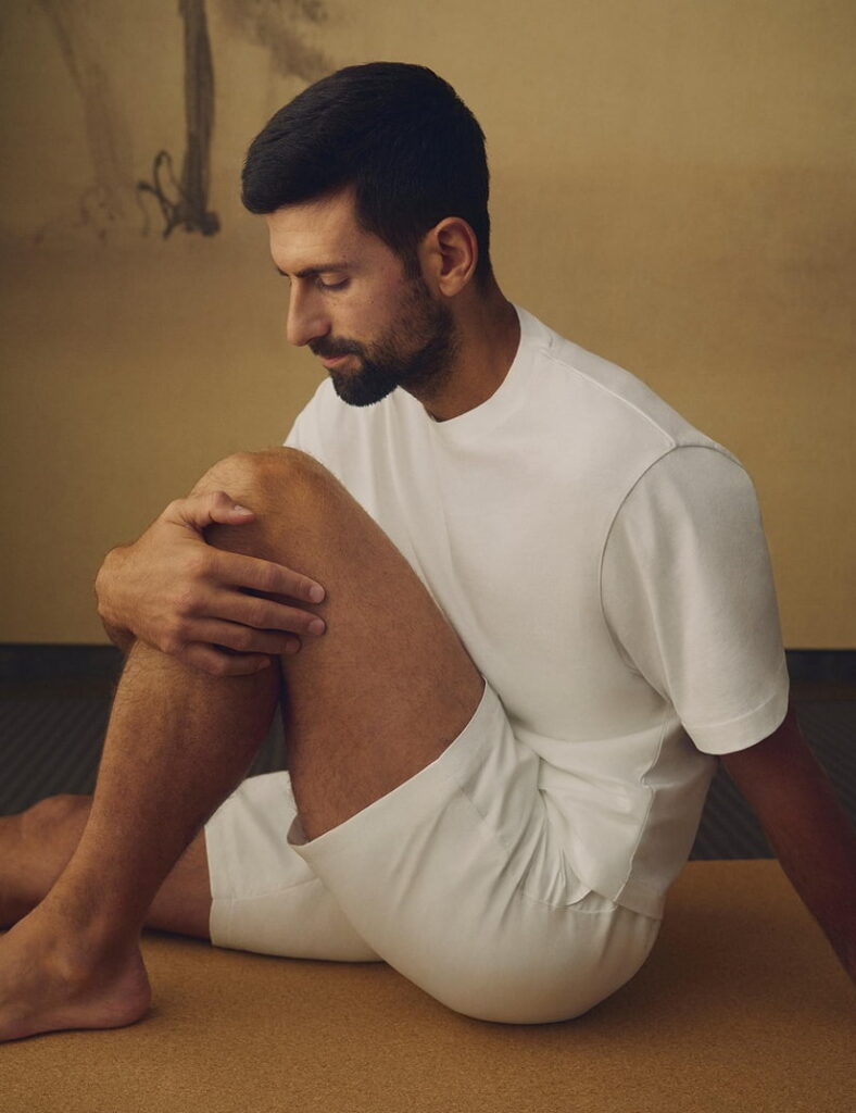Novak Djokovic acostado de espaldas sobre una superficie de madera, mostrando una flexibilidad extrema al llevar su pierna totalmente estirada hacia su cabeza, usando un polo Lacoste terracota.