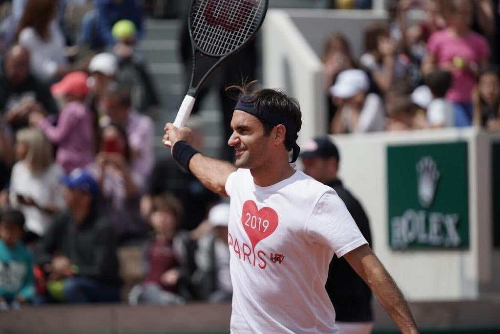 Roger Federer sonriendo en una cancha de tenis, sosteniendo su raqueta Wilson Pro Staff de aro ancho durante un entrenamiento.